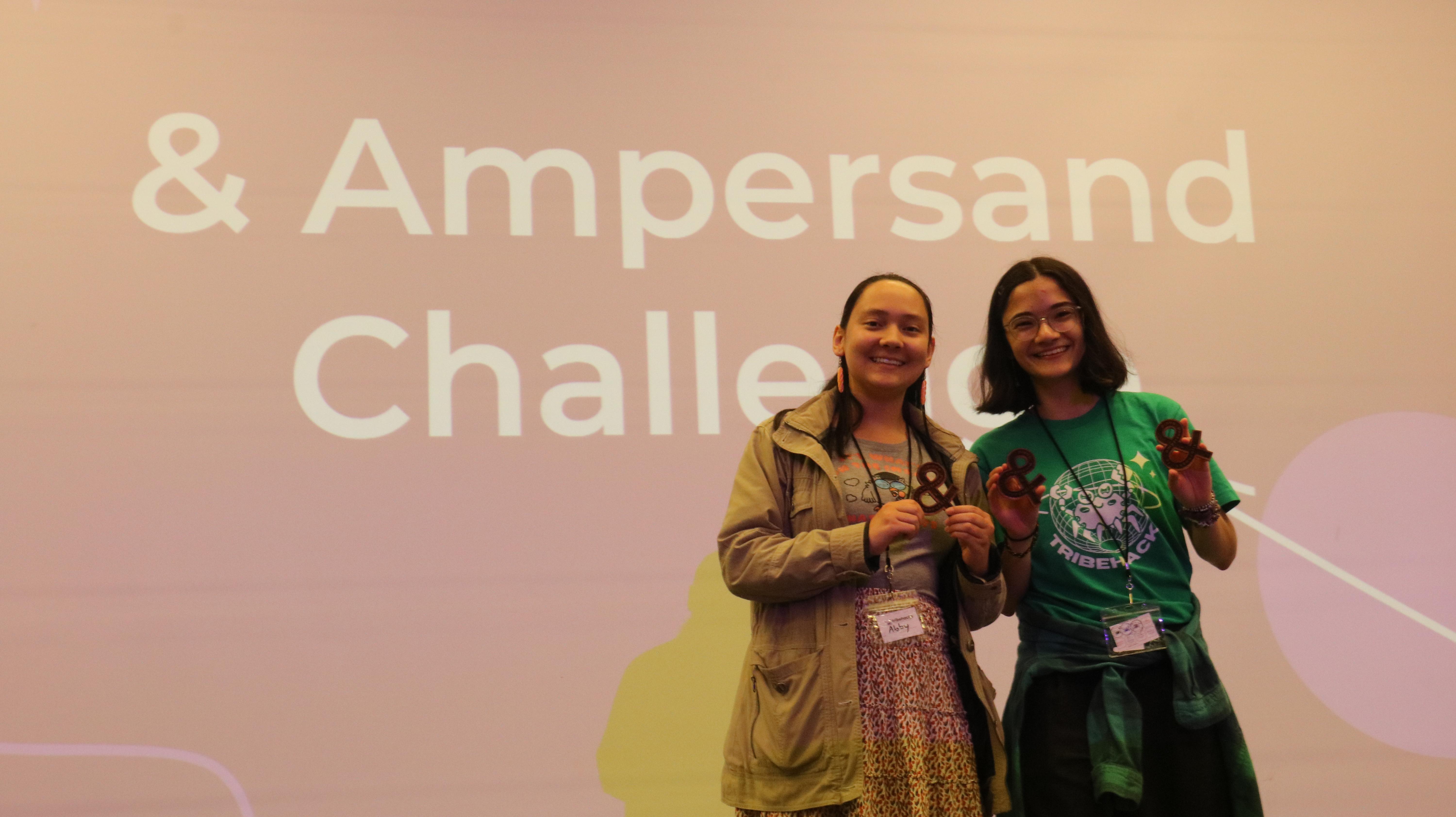 Two people smiling and holding wooden ampersands. They are standing in front of a whiteboard that says 'Ampersand Challenge'